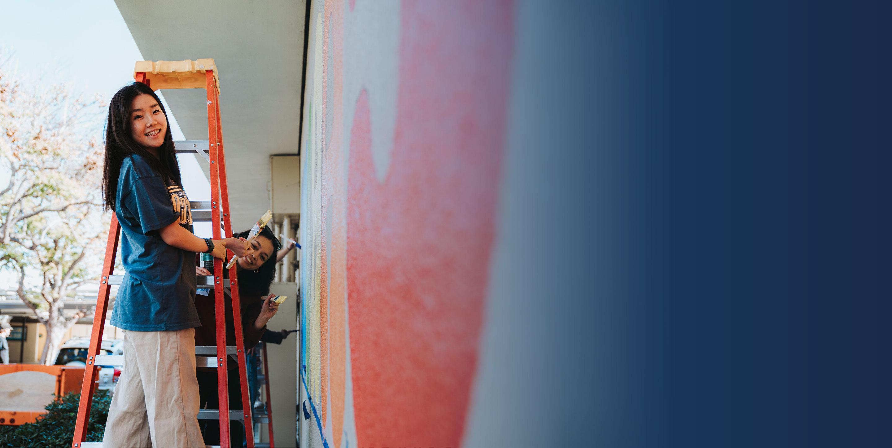 Students stand on ladder with paintbrushes next to wall mural